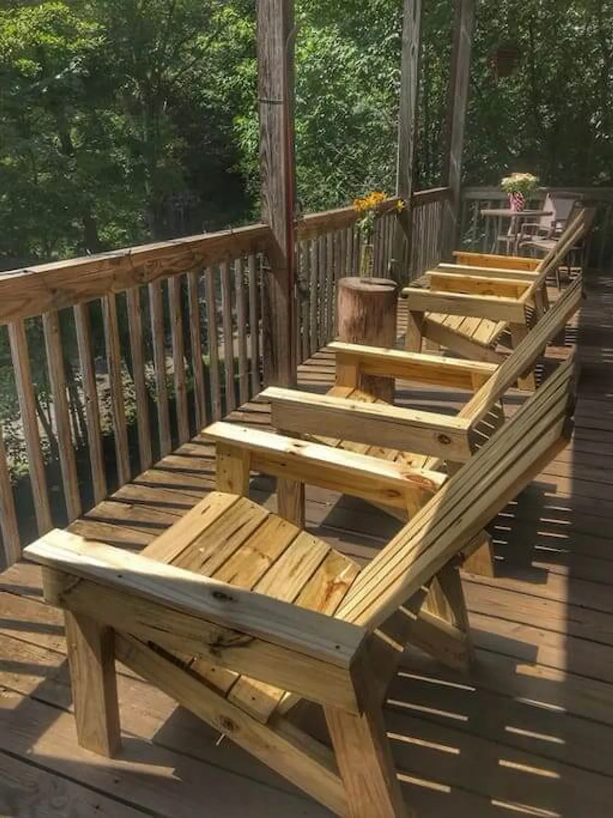 This image captures a close-up of a deck outfitted with rustic wooden chairs, a stump table, and a flower vase. The wooden railing frames the view of nature beyond, while the warm lighting suggests either morning or evening, evoking a sense of relaxation and connection with the outdoors.