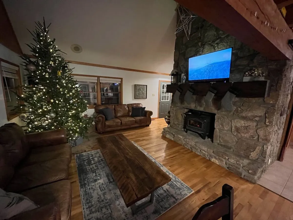 A warm and festive living room featuring a stone fireplace, leather sofas, a wooden coffee table, and a decorated Christmas tree. The space exudes a cozy atmosphere, perfect for gatherings by the fire or movie nights after a day on the slopes.