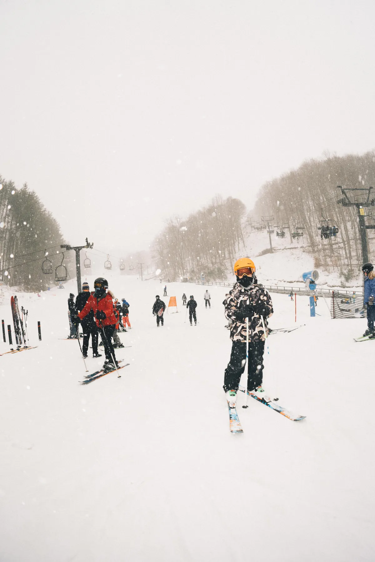 A skier stands in the foreground with a ski lift and a group of people skiing in the background, all set against a snow-covered landscape. The scene buzzes with energy and excitement, capturing people actively enjoying the slopes and the resort environment.