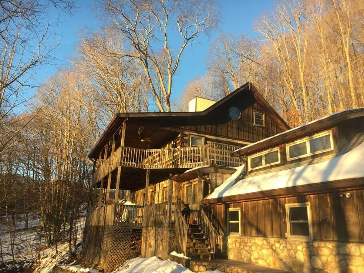 An exterior view of a wooden balcony attached to a stone-clad house, overlooking a snow-covered ground and surrounded by tall trees. The scene conveys both the privacy and the expansive outdoor living experience, with ample space for gathering and enjoying nature.
