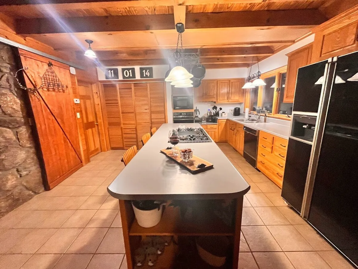 A bright, inviting kitchen featuring a prominent island, sleek wooden cabinets, dining chairs, and a set of wine glasses. The scene suggests a space ideal for both cooking and entertaining, with generous counter space and a layout that fosters social interaction. The airy design ensures the chef remains connected to guests or family.
