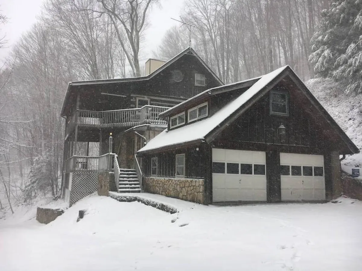 The image shows a snow-covered house nestled in the mountains, featuring a large wooden deck with outdoor seating. The deck is surrounded by a tranquil forested backdrop, and footprints in the snow suggest recent outdoor activity. The inviting deck space hints at cozy gatherings and relaxation, with the mountains visible beyond the trees.