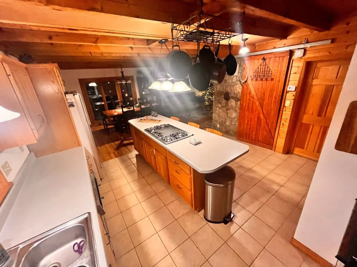 A kitchen and dining area with an expansive island, hanging pots, wooden beams, and a textured stone wall. The design merges rustic charm with modern functionality, providing ample space for cooking, entertaining, and family gatherings. The sight-lines between kitchen and dining area enhance communal activities and conversations.