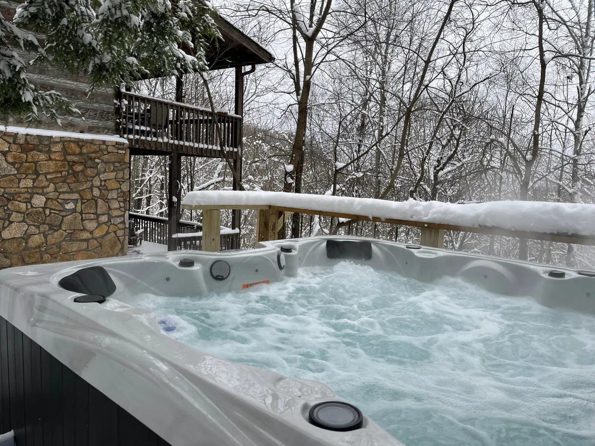 A serene exterior scene featuring a steaming hot tub on a deck surrounded by snow-covered trees and a sturdy wooden railing. The atmosphere is tranquil, suggesting a perfect spot for après-ski relaxation with friends as you watch the snow fall.