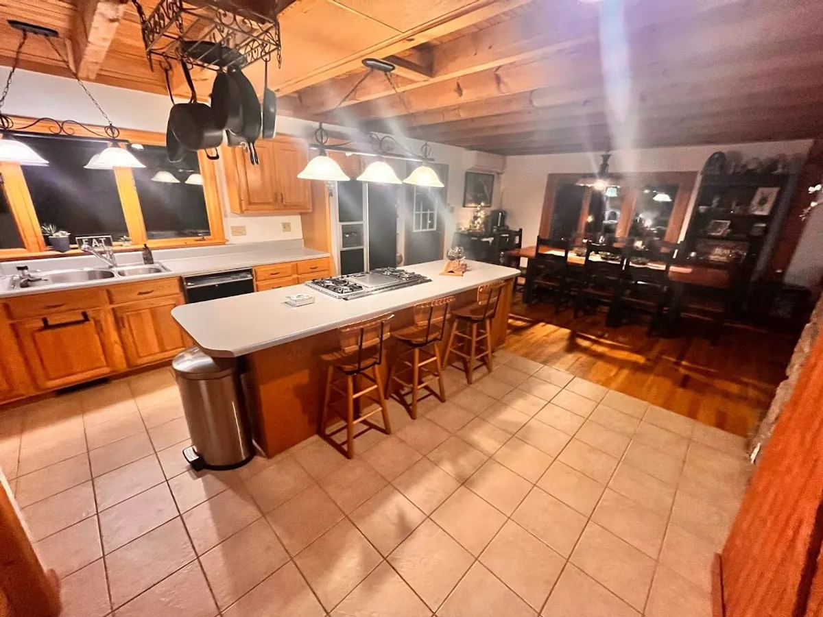 An open kitchen and dining area with an island counter and stools, kitchen appliances, dining table with chairs, and large windows that let in plenty of natural light. The scene evokes a sense of comfort and togetherness, with the kitchen as a social hub.