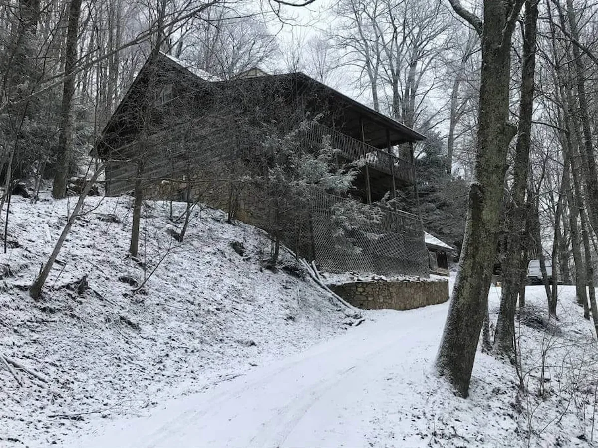 A rustic wooden house surrounded by snow, featuring a snow-covered path and flanked by trees. The peaceful environment suggests opportunities for snowball fights, building snowmen in the yard, and gathering on the porch for stargazing or cocoa.