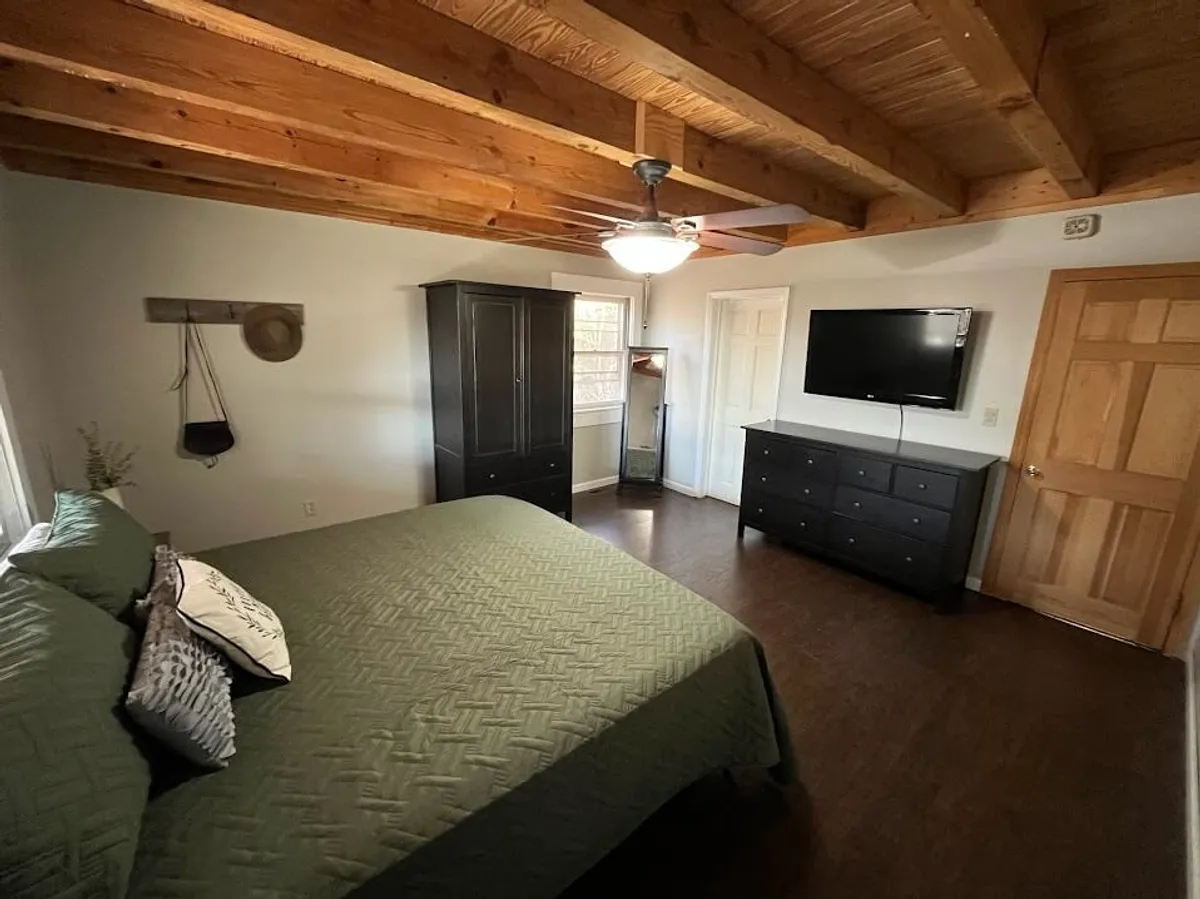 A cozy bedroom with a bed covered in a green quilt, a dark wooden dresser, a wall-mounted TV, and exposed wooden beams on the ceiling. The space exudes comfort and warmth, inviting relaxation after a day of mountain adventures.